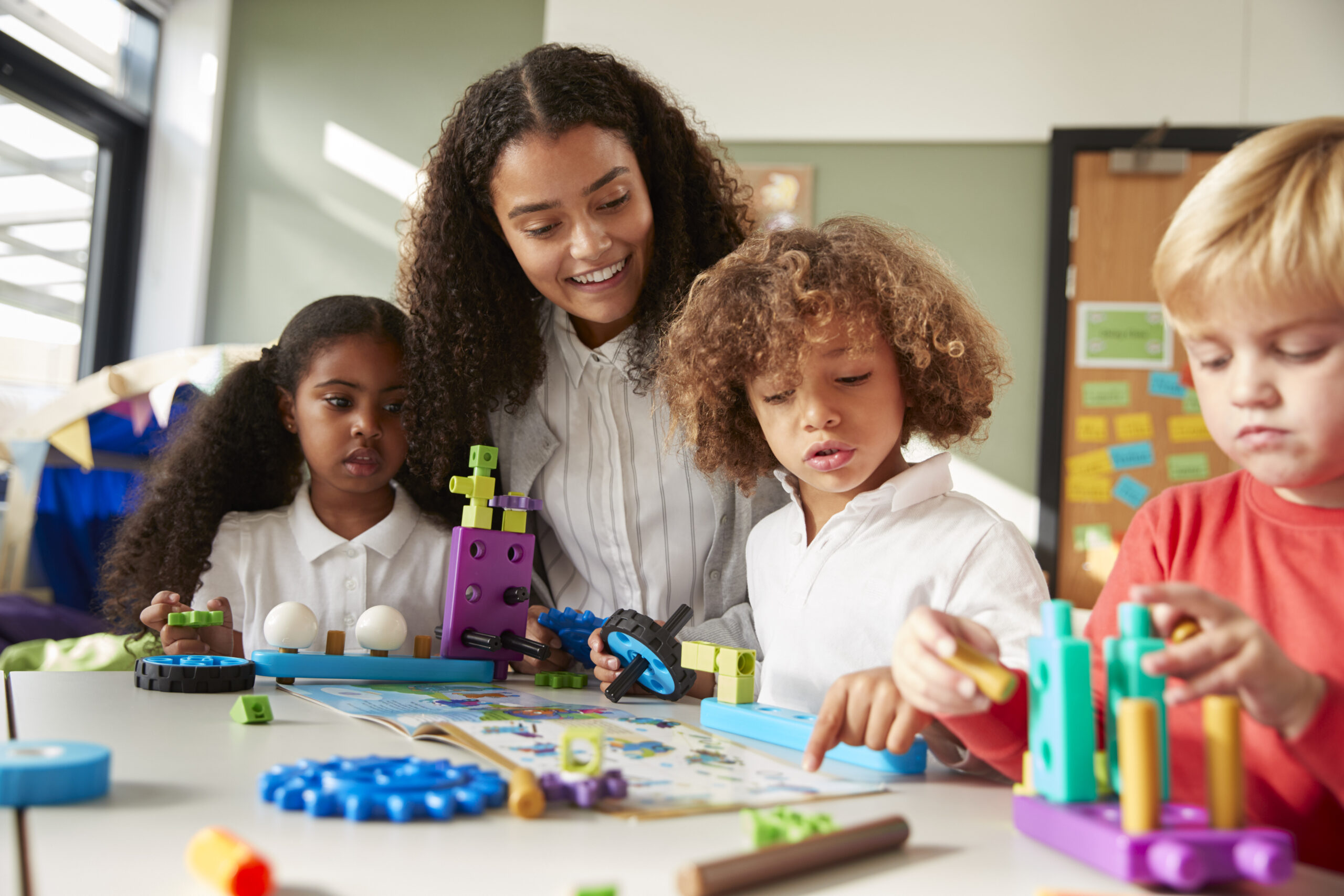 Female teacher sitting at table in play room with three kindergartne children constructing, selective focus
